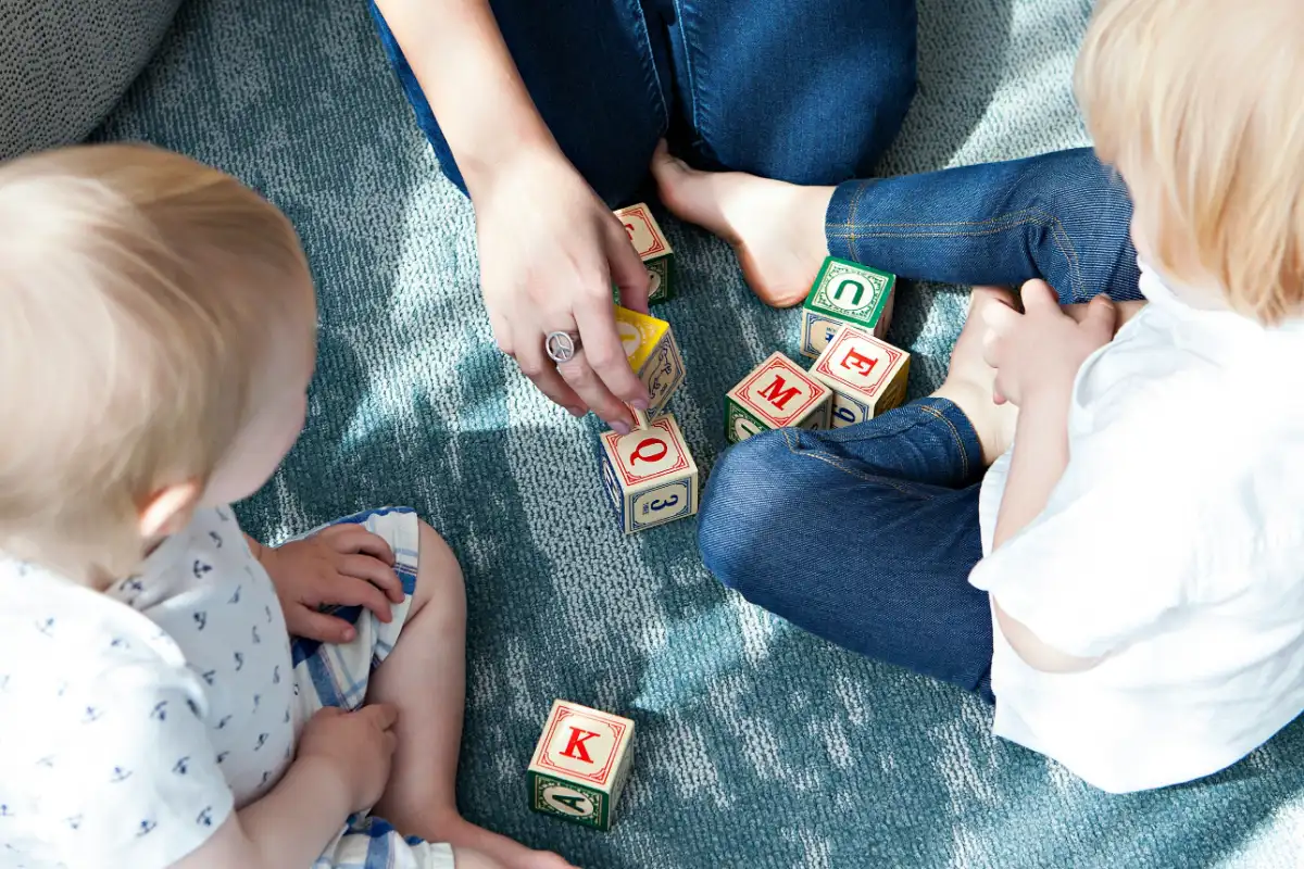 Une adulte assise par terre avec deux jeunes enfants, qui empilent des cubes en bois.